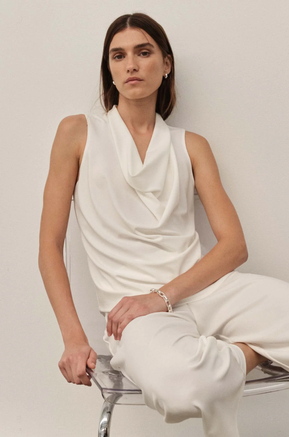 Woman wearing a white sleeveless top and pants sitting on a chair against a plain background