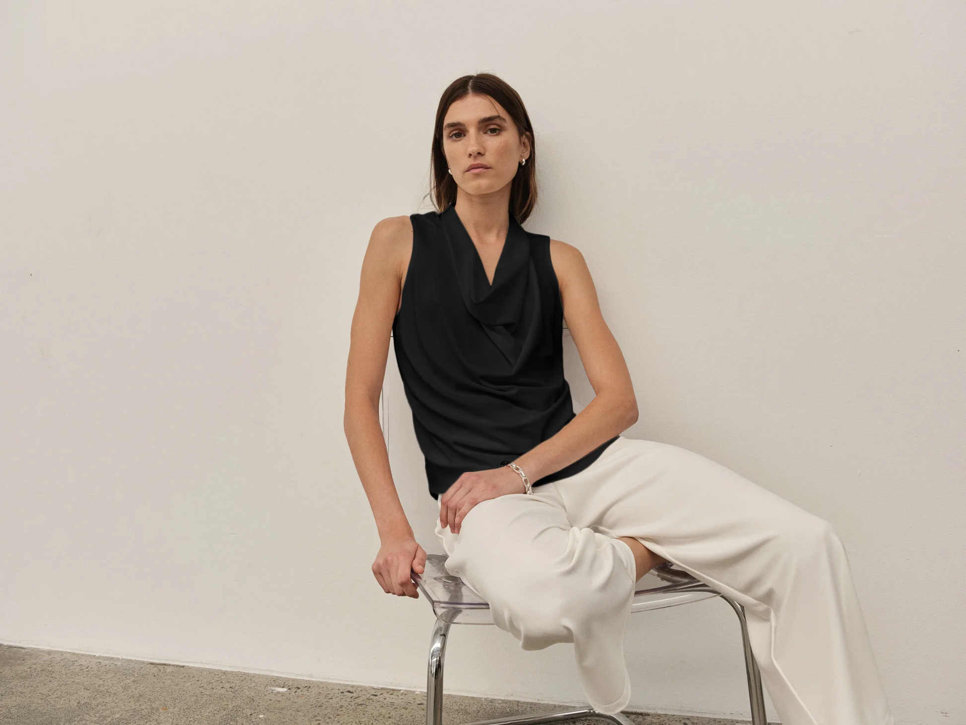 Woman sitting on a chair wearing a black sleeveless top and white pants against a plain background