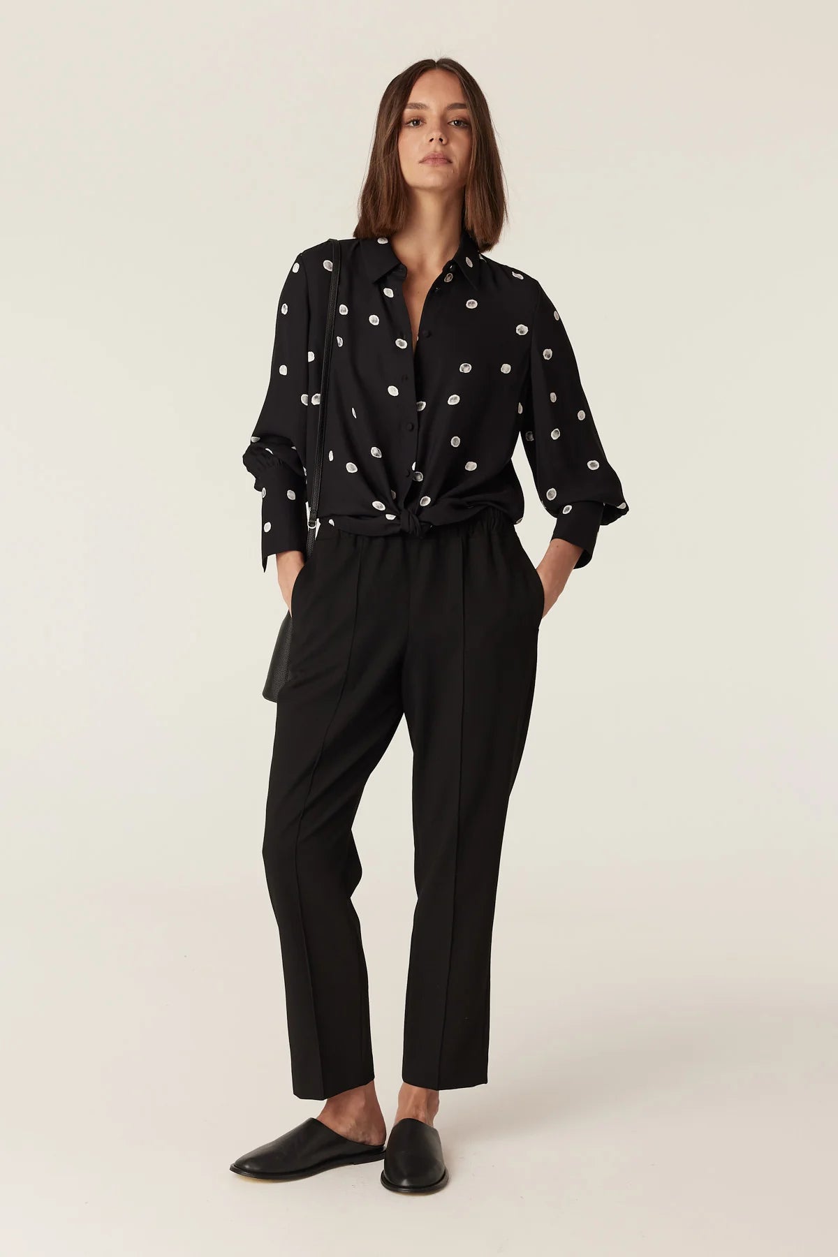 Woman wearing the Cable Melbourne Hepburn Blouse in Black Spot, tied at the hem over black trousers