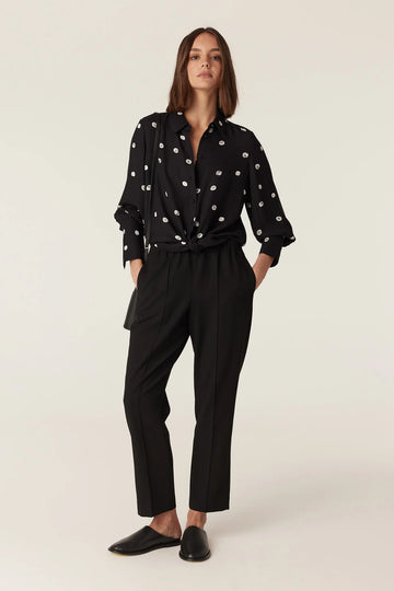 Woman wearing the Cable Melbourne Hepburn Blouse in Black Spot, tied at the hem over black trousers